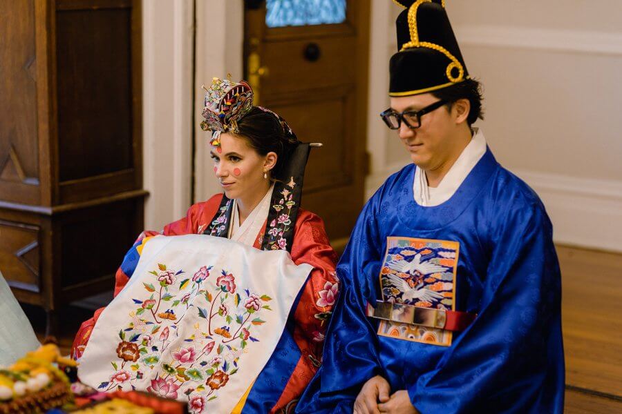 Bride and groom in traditional Korean wedding outfits sit at a tea ceremony