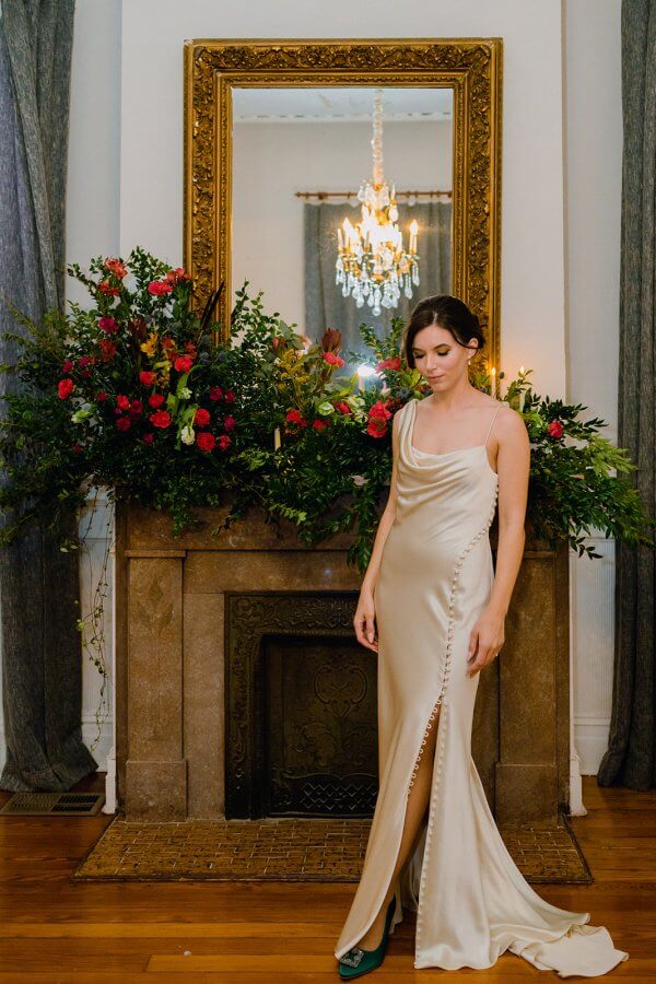 Bride in glamorous cream gown stands in front of a flower display and firepalce