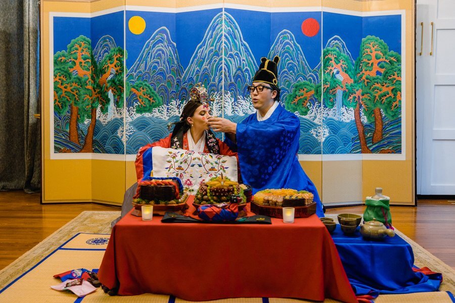 Bride and groom in traditional Korean wedding outfits sit at a tea ceremony in front a lot of colorful food