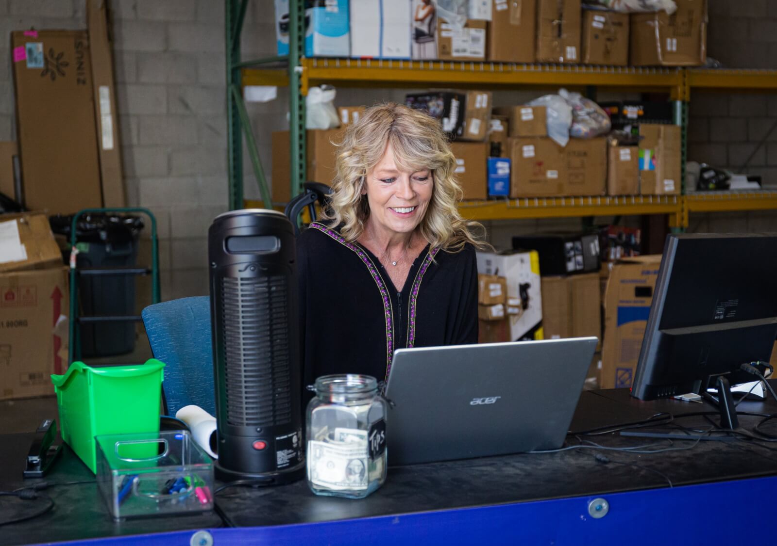 Blonde woman in a black top typing on a laptop in a warehouse.