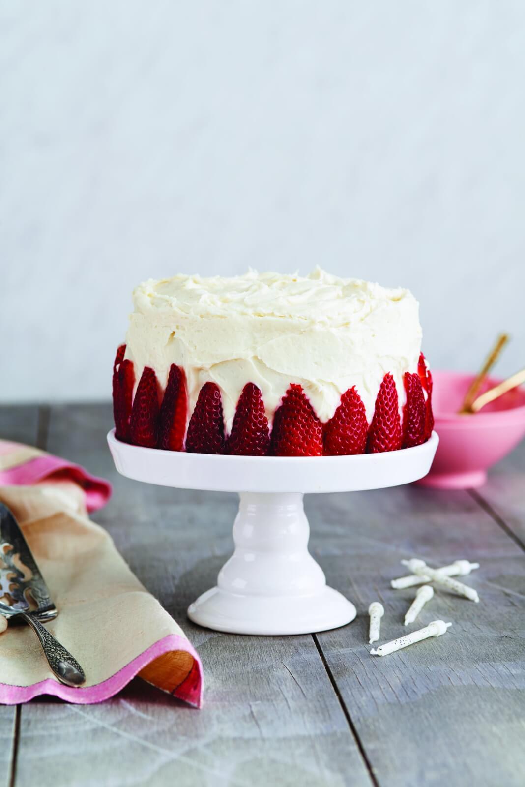 Small white cake with sliced strawberries on the side, sitting on a white cakestand.